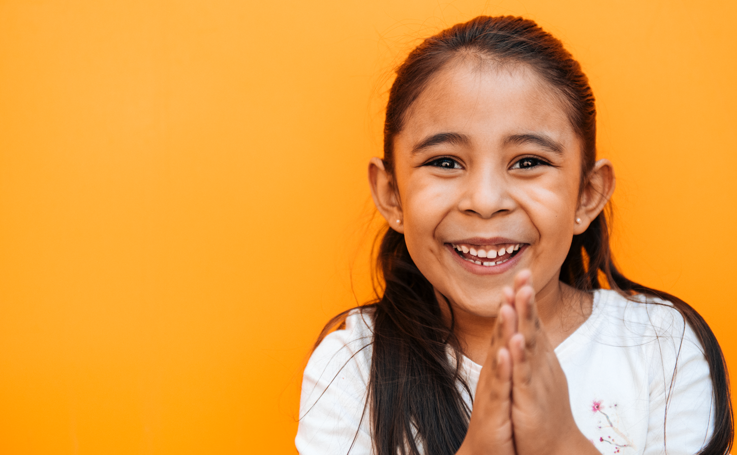 A little girl grinning widely and clapping her hands, on a bright yellow-orange background