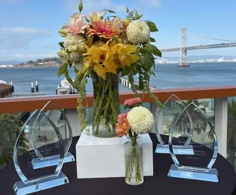 A small table with our 2025 teardrop-shaped pro bono awards plaques on it, next to two vases of orange, yellow, and white flowers, with the Bay Bridge under a blue sky in the background