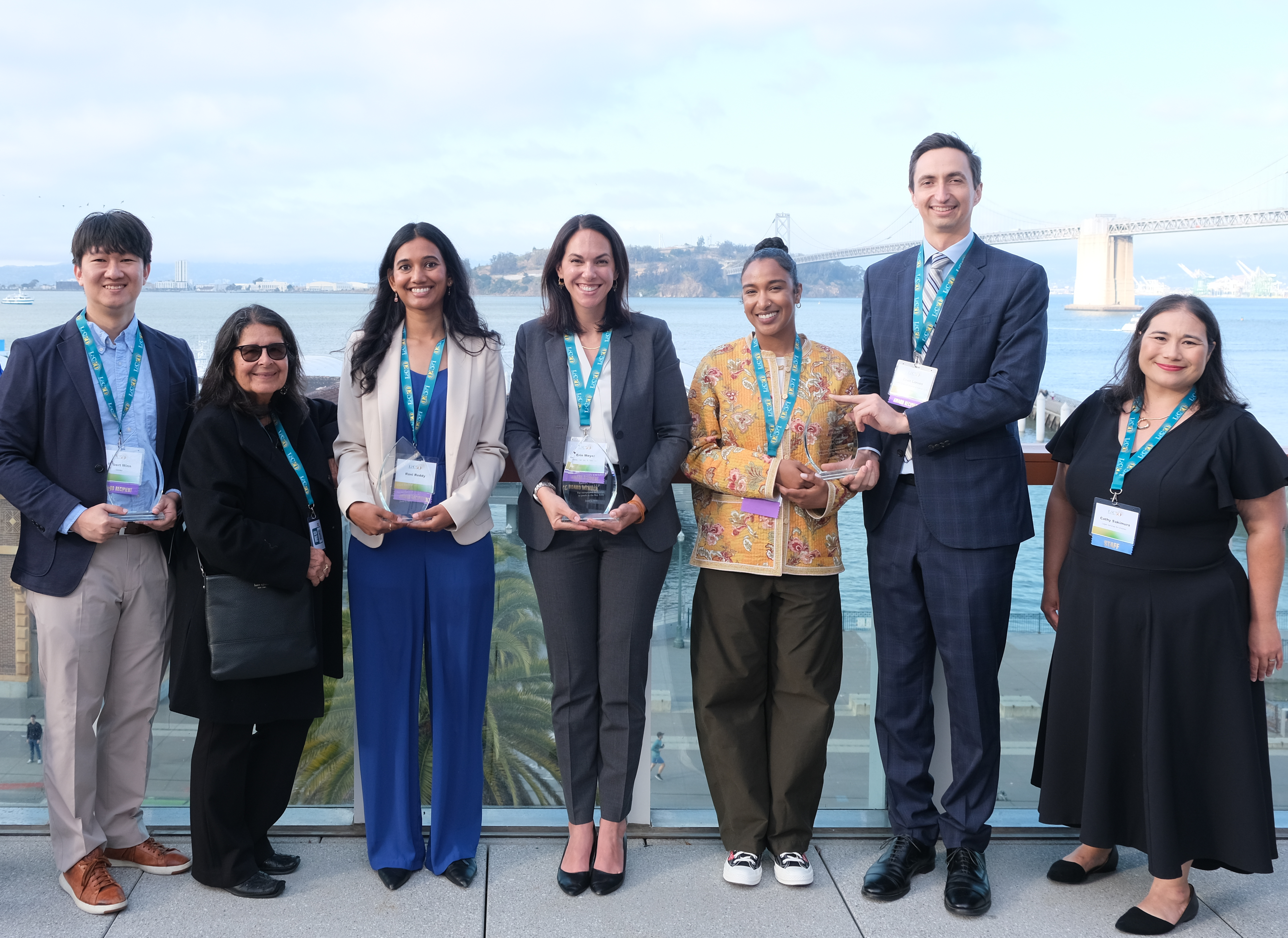 All of LSC's 2025 pro bono awardees with their awards plaques, alongside LSC's Executive Director Cathy Sakimura and Trinidad Madrigal, former LSC volunteer for whom our Trinidad Madrigal Excellence in Child-Centered Representation Award is named. They stand on a rooftop terrace with the Bay Bridge in the background.