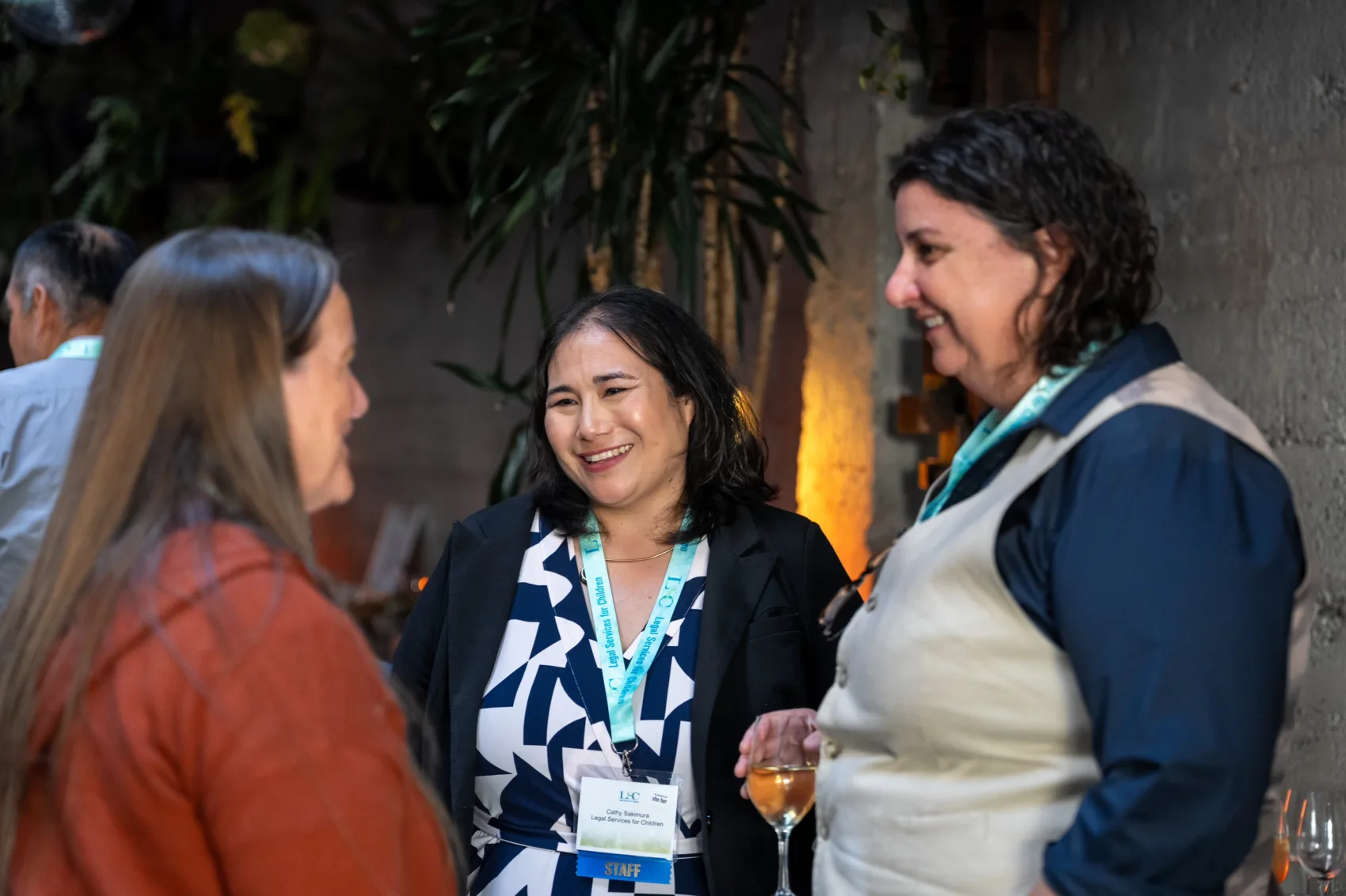 Three people smiling and chatting in front of some indoor trees. From left, Excellence in Service to Youth awardee Perryn Reis, LSC Executive Director Cathy Sakimura, and LSC Senior Staff Attorney Jen Daly