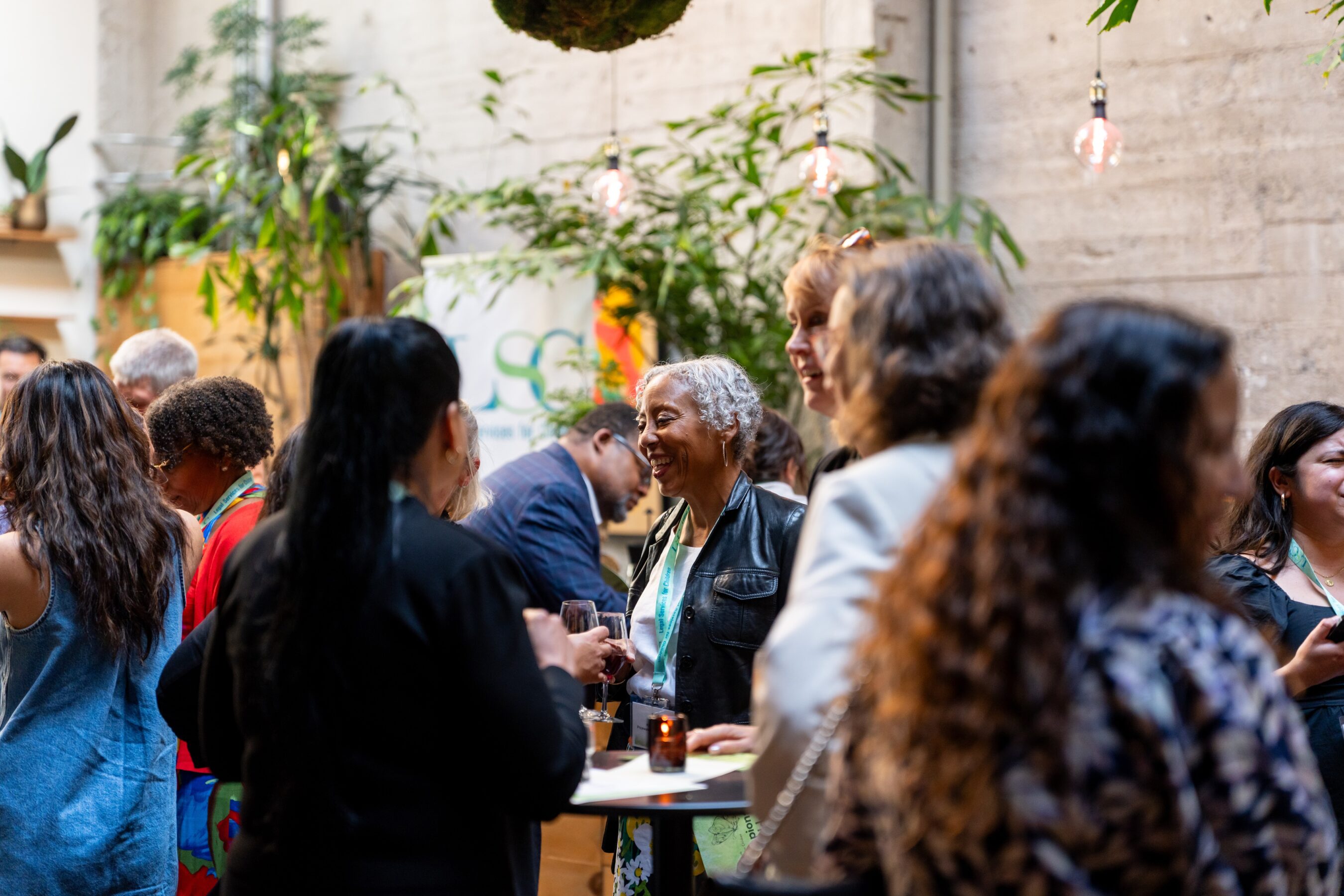 A crowd of event attendees stand around tables smiling and chatting in front of a stone wall lined with indoor trees