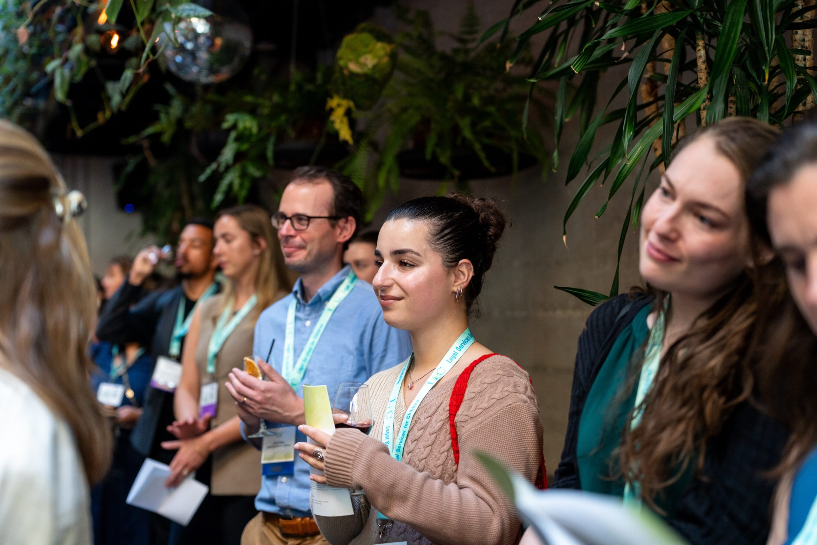 A crowd of event attendees stand smiling and clapping in front of a wall lined with indoor trees