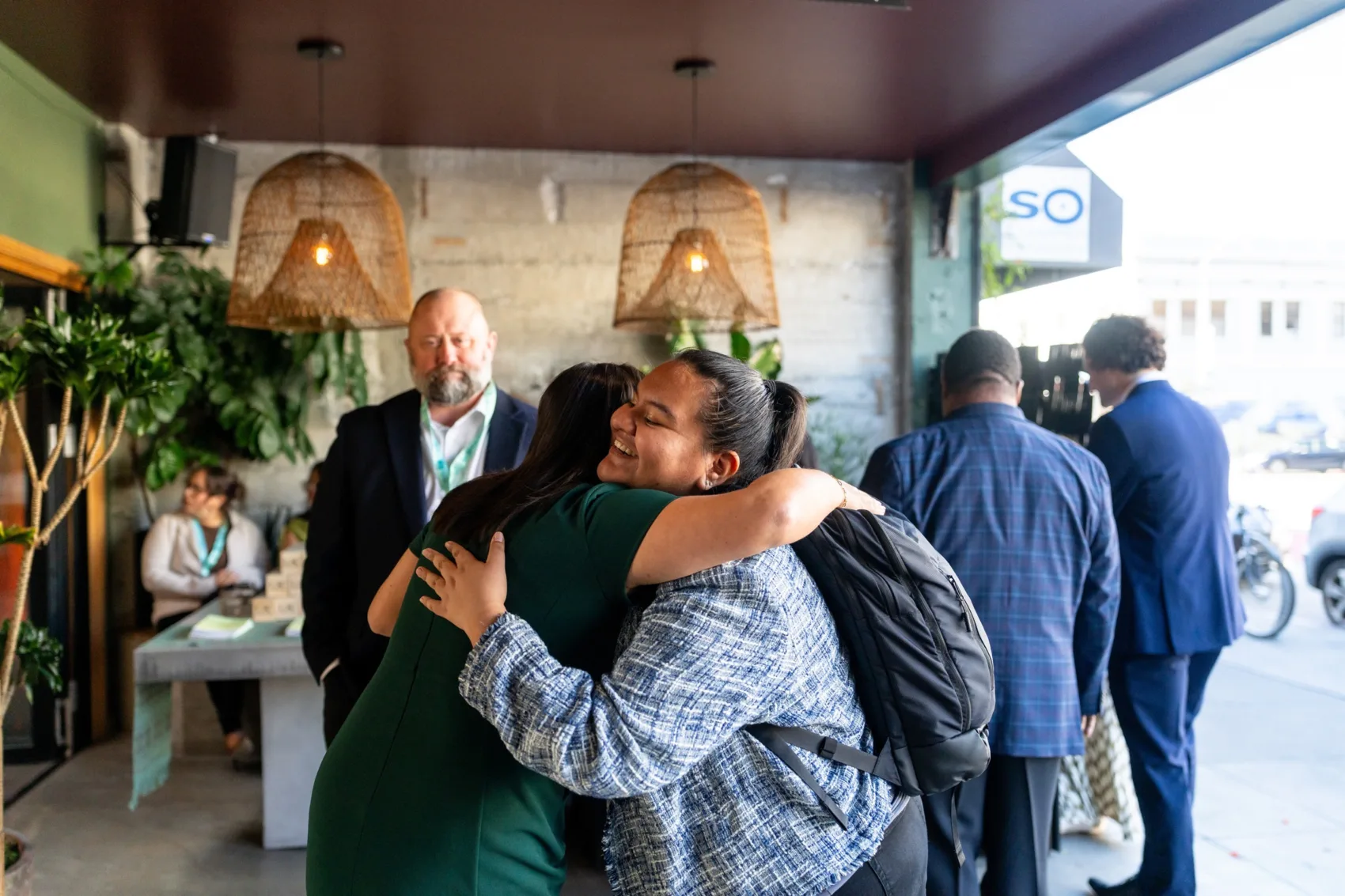 Two people hugging in an open foyer area with other people entering the space in the background