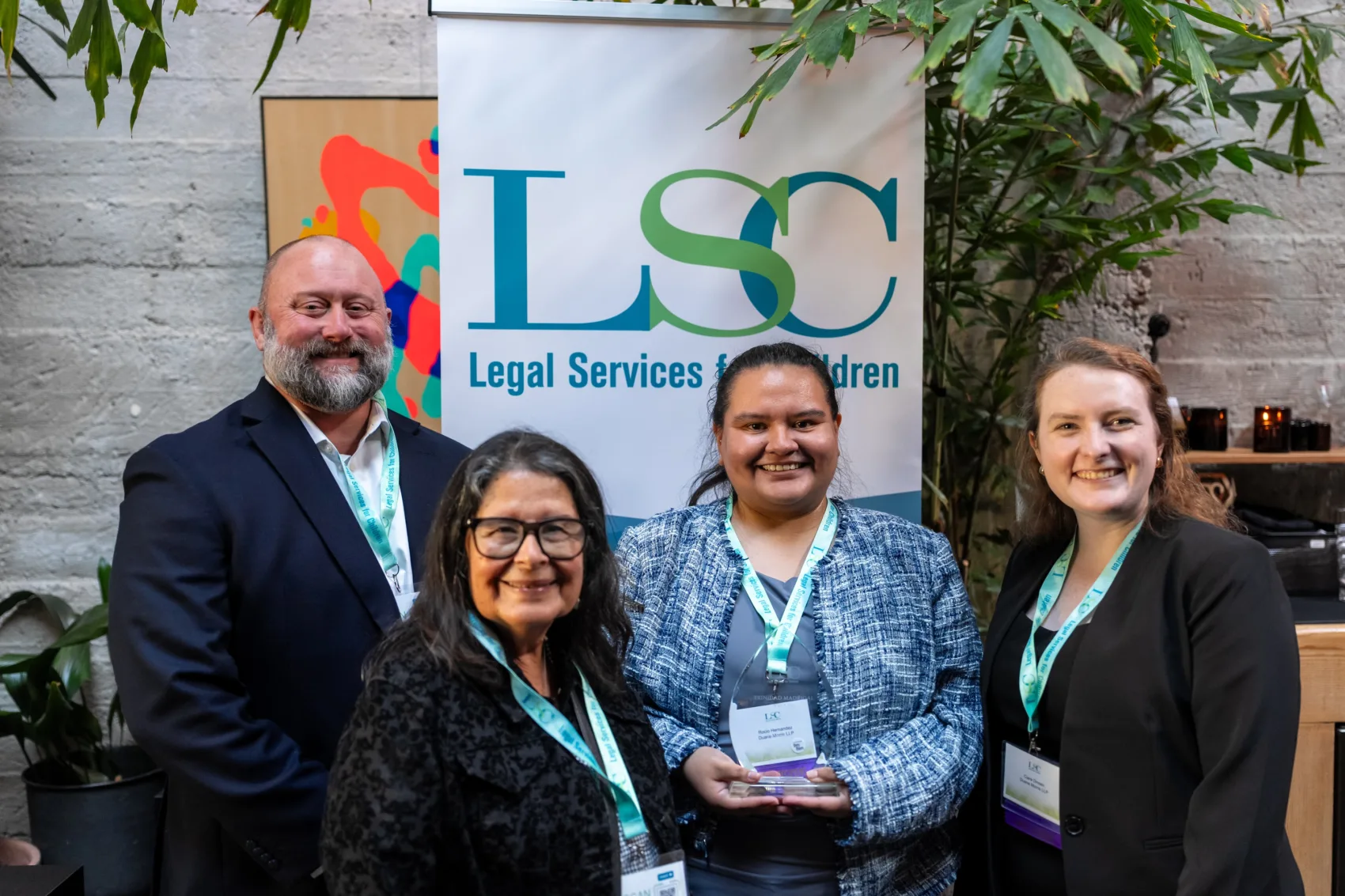 Three LSC Excellence in Pro Bono awardees standing with their award alongside former LSC volunteer Trinidad Madrigal, in front of an LSC banner