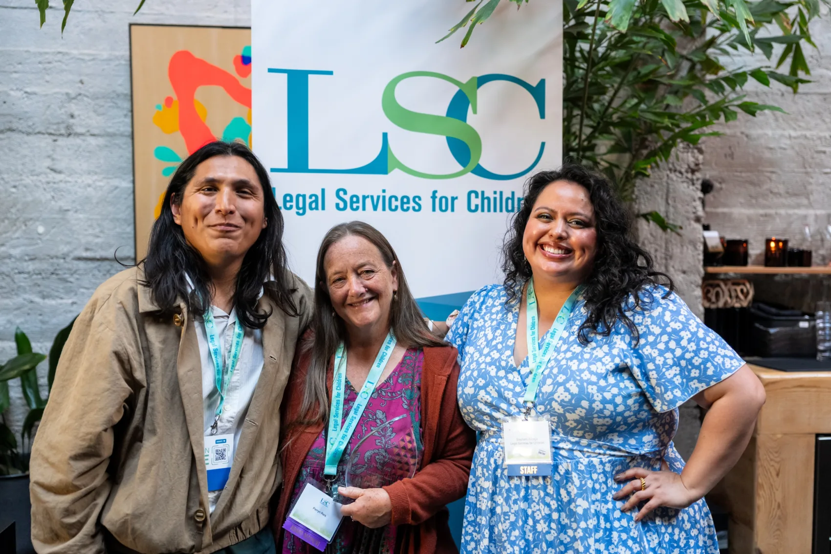 LSC Excellence in Service to Youth awardee Perryn Reis standing with LSC social worker Fernando Antunez and LSC Associate Legal Director Stephany Arzaga in front of an LSC banner