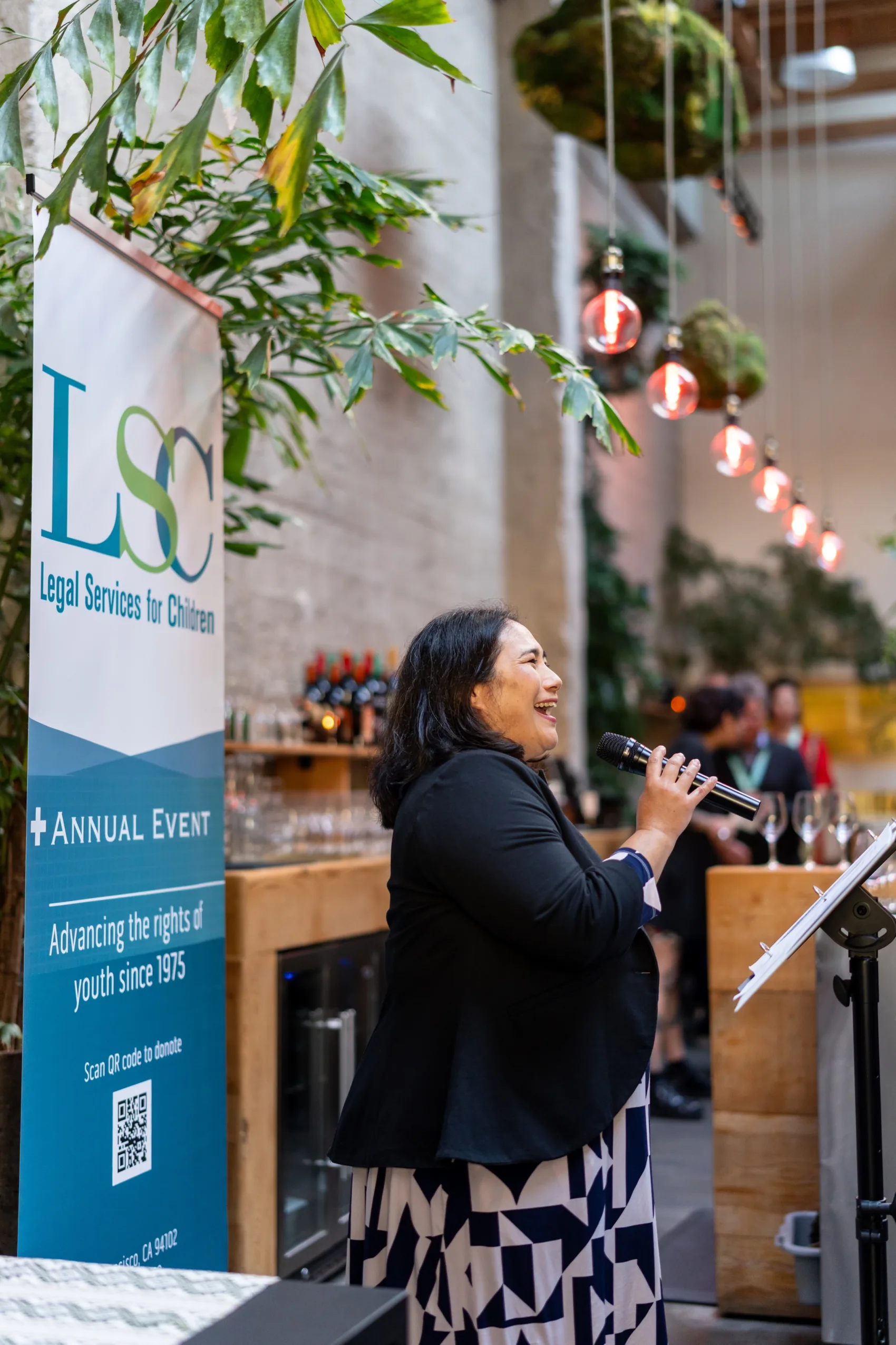 LSC's Executive Director Cathy Sakimura stands smiling and holding a microphone in front of an LSC banner as she addresses attendees at LSC's 2026 annual event